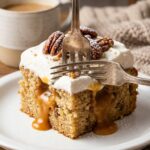 A close-up of a fork cutting into a slice of poke cake, with caramel oozing out of the cake's holes, topped with pecans.