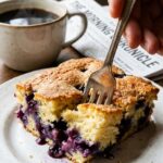 A close-up of a fork cutting into a slice of tender blueberry cake, showing the moist crumb and jammy berries, with a coffee mug in the background.