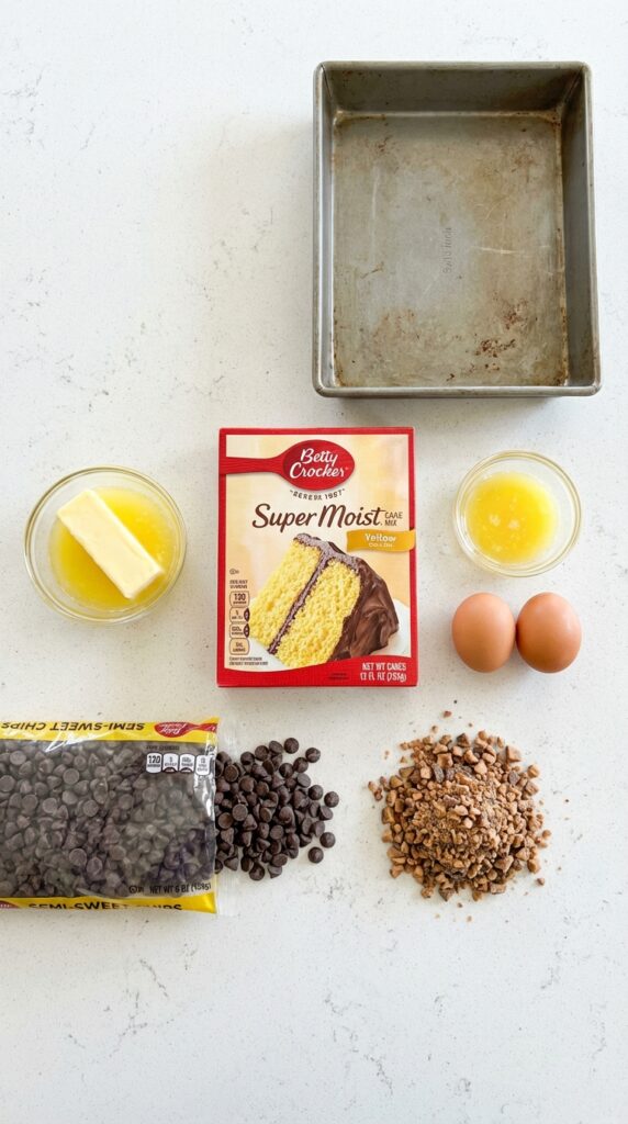 A top-down photograph displaying a yellow cake mix box, butter, eggs, chocolate chips, and toffee bits on a kitchen counter.