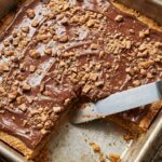 An angled view looking into a metal baking pan filled with baked golden toffee bars covered in melted chocolate and crunchy toffee bits, with a spatula lifting a corner piece.
