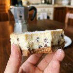 A close-up of a hand holding a cannoli cheesecake bar with a bite taken out, showing the creamy interior and chocolate chips.