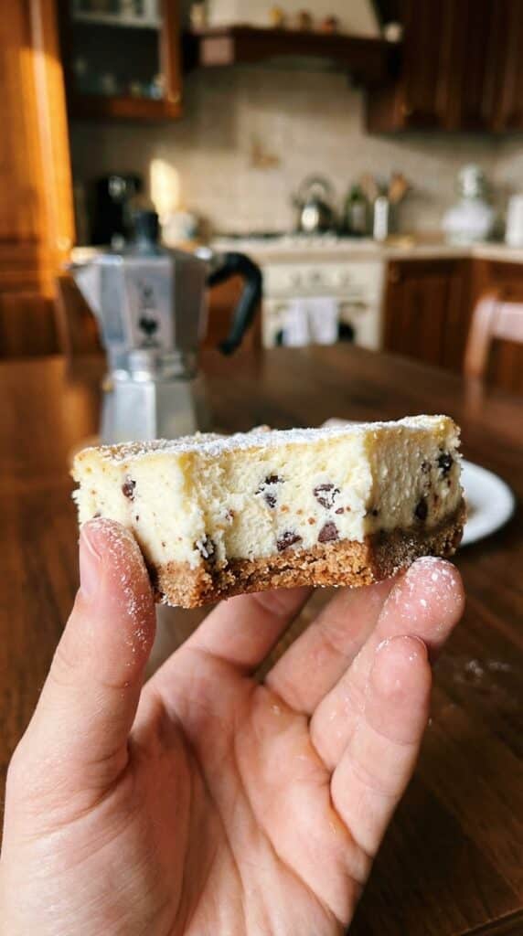 A close-up of a hand holding a cannoli cheesecake bar with a bite taken out, showing the creamy interior and chocolate chips.