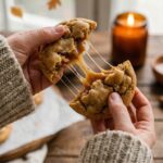 Two hands in a chunky knit sweater breaking apart a warm caramel-stuffed apple cookie.
