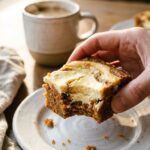 A close-up of a hand holding a swirled carrot cake bar with a bite taken out, showing the moist interior, with coffee in the background.