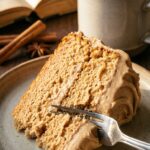 A close-up of a fork cutting into a slice of moist chai cake with thick frosting, with a mug of tea in the background.