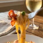 A close-up of a silver fork twirling creamy spaghetti with bacon and peas, with a glass of white wine in the background.