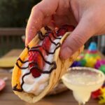 A close-up of a hand holding a loaded strawberry cheesecake taco with a party setting in the blurred background.