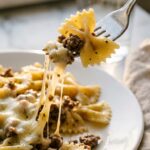 A close-up of a fork lifting a piece of bowtie pasta and ground beef from a plate, showing a long string of melted cheese.