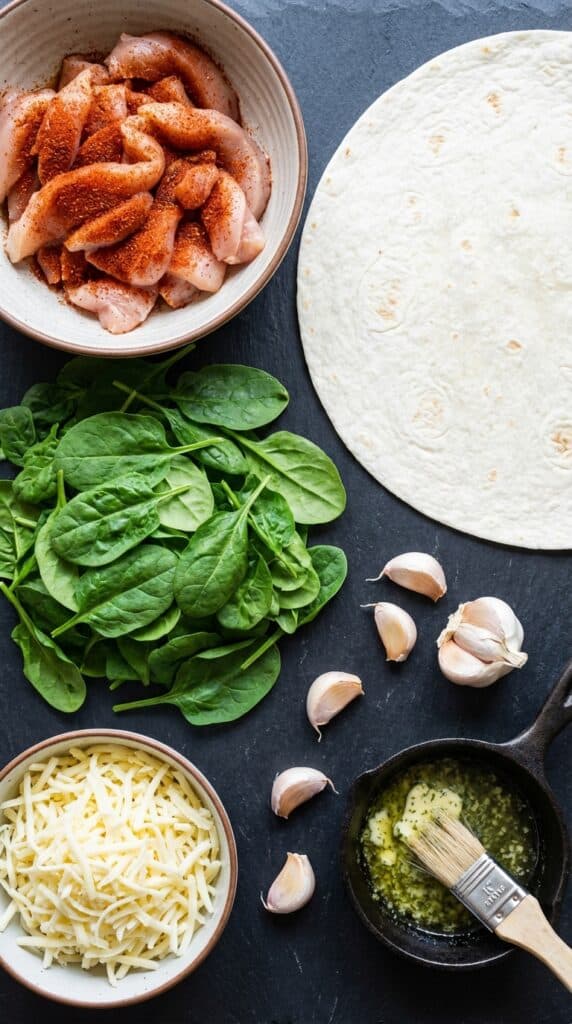 A flat lay showing seasoned raw chicken, a flour tortilla, shredded cheese, fresh spinach, garlic, and melted butter on a slate board.