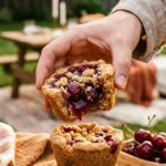 A close-up of a hand holding a cherry pie cookie cup with a bite taken out, showing the fruit filling, in a backyard setting.