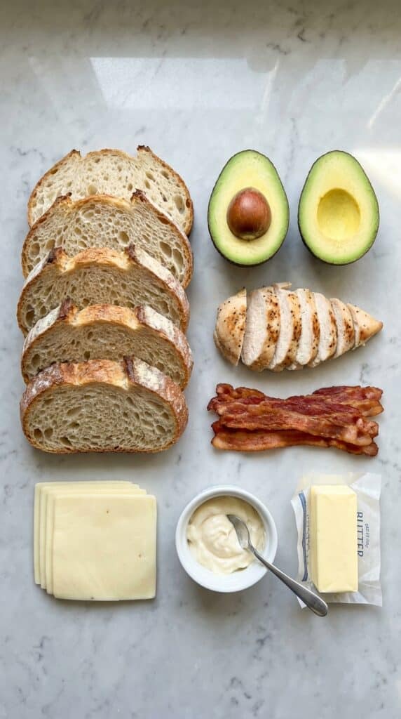 A flat lay showing sourdough bread, avocado, sliced chicken, bacon, provolone cheese, and mayo on a marble surface.