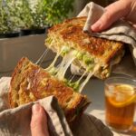 Two hands pulling apart a hot toasted chicken and avocado sandwich, creating a long stringy cheese pull.