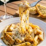 A close-up action shot of a fork lifting a bite of creamy chicken pasta, creating a massive stringy mozzarella cheese pull.