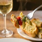 A close-up of a fork holding a piece of pesto-coated pasta, chicken, and a cherry tomato.