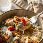 A close-up of a fork lifting a piece of chicken, tomato, and rice coated in creamy garlic sauce from a colorful bowl.