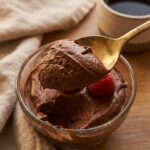 A close-up of a gold spoon lifting a thick, smooth dollop of dark chocolate mousse with a raspberry on top.