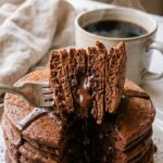A close-up of a fork cutting into a stack of chocolate pancakes, revealing a fluffy interior and melted chocolate chips.