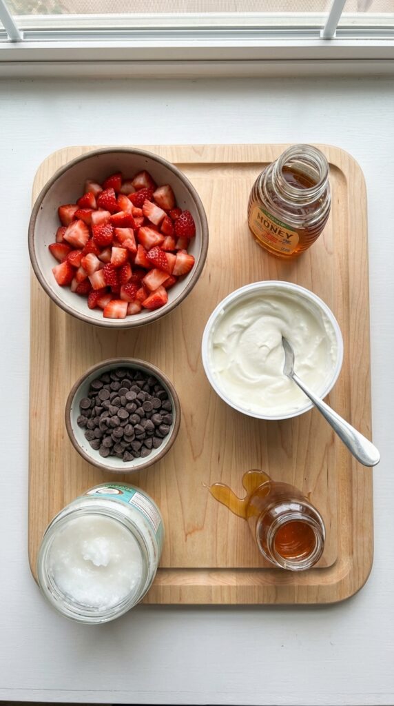 A flat lay showing diced strawberries, Greek yogurt, dark chocolate chips, coconut oil, and honey on a wooden board.