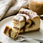 A close-up of a slice of cinnamon roll cheesecake on a plate, showing the gooey brown sugar swirl inside, with a fork taking a bite.