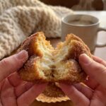 A close-up of hands pulling apart a warm cinnamon sugar muffin to reveal the cream cheese center, with sugar dusting the fingers.