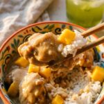 A close-up of chopsticks lifting a piece of saucy coconut chicken, rice, and mango from a bowl.