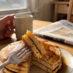 A close-up of a fork cutting into a stack of coffee pancakes, revealing a fluffy interior with espresso syrup dripping down. Social