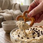 A close-up of a hand holding a pretzel scooped full of chocolate chip cookie dough dip.