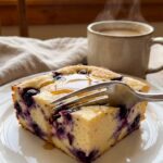 A close-up of a fork cutting into a thick slice of creamy blueberry cottage cheese bake on a plate, with coffee in the background.