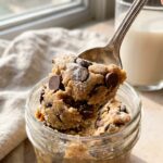 A close-up of a spoon lifting a thick scoop of chocolate chip cookie dough from a jar.