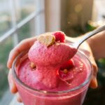 A close-up of a spoon lifting a thick, perfectly smooth scoop of pink raspberry mousse from a bowl.