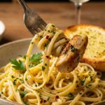 A close-up of a fork twirling glossy, herb-coated linguine and a piece of chicken from a bowl.
