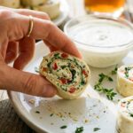 A close-up of a hand holding a single cream cheese and bacon tortilla bite, with ranch dip in the background.