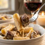 A close-up of a fork lifting a piece of rigatoni pasta and a seared steak bite, dripping with creamy parmesan sauce.