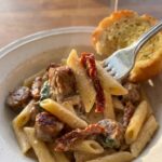 A close-up of a fork lifting a bite of creamy sausage pasta with a sun-dried tomato from a bowl, with wine in the background.