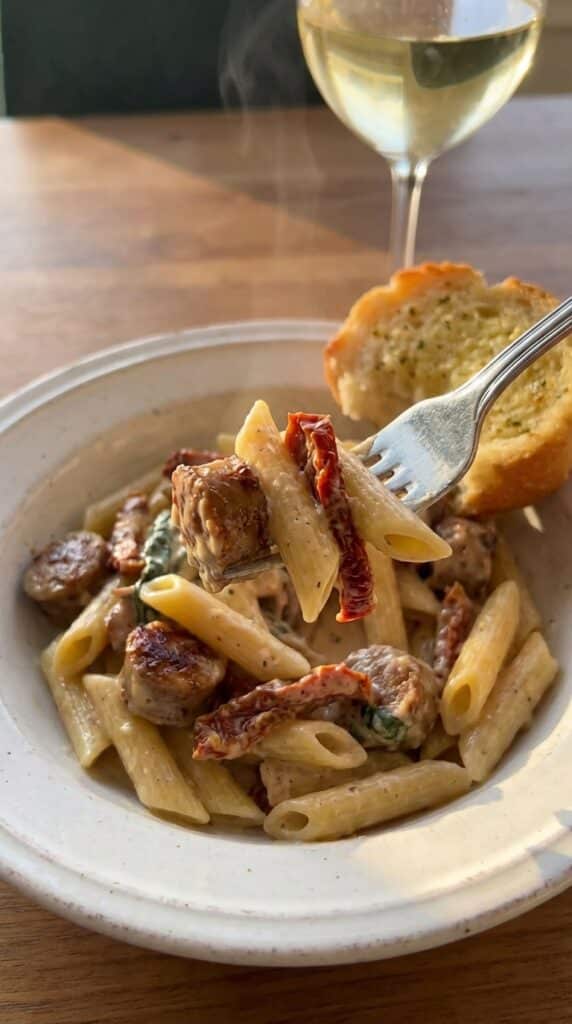 A close-up of a fork lifting a bite of creamy sausage pasta with a sun-dried tomato from a bowl, with wine in the background.