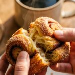 Hands pulling apart a warm cinnamon sugar cruffin, showing the soft, flaky, buttery layers inside with steam rising.