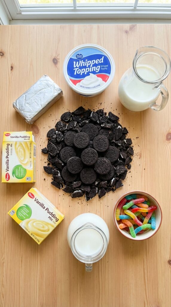A flat lay showing chocolate sandwich cookies, cream cheese, vanilla pudding boxes, whipped topping, milk, and gummy worms on a wooden board.