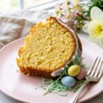 A close-up of a slice of vanilla bundt cake on a pink plate, with white glaze, green coconut, and pastel candy eggs next to it.