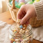 A hand reaching into a glass jar to grab a handful of Easter Chex mix with a blurred Easter basket in the background.