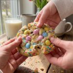 A close-up of two hands breaking a thick Easter cookie in half, showing a soft, gooey center with melted pastel candies.