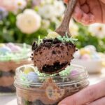 A close-up shot of a hand using a wooden spoon to scoop a layered Easter Dirt Cup dessert topped with Peeps and candy.