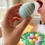A close-up of a hand holding a light blue, chocolate-covered Oreo egg treat, with an Easter basket in the background.