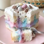 A close-up side view of a slice of Easter poke cake on a plate, showing the pastel pudding stripes inside the white cake.