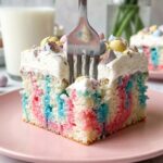 A close-up of a fork cutting into a slice of colorful Easter poke cake with whipped cream and candy eggs.