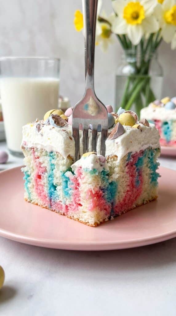 A close-up of a fork cutting into a slice of colorful Easter poke cake with whipped cream and candy eggs.