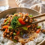 A close-up of chopsticks lifting a piece of teriyaki ground turkey and broccoli from a rice bowl, with steam rising.