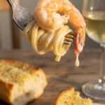 A close-up of a fork holding linguine pasta and a creamy garlic shrimp, with the sauce dripping slightly, and wine in the background.