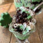 A close-up of a large spoon scooping out a messy, delicious serving of layered brownie, green pudding, and whipped cream