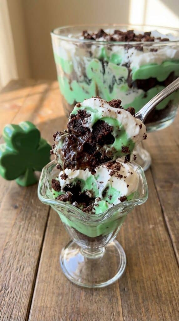 A close-up of a large spoon scooping out a messy, delicious serving of layered brownie, green pudding, and whipped cream