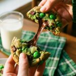 Hands pulling apart a warm green St. Patrick's Day cookie, showing melted chocolate and a soft center.